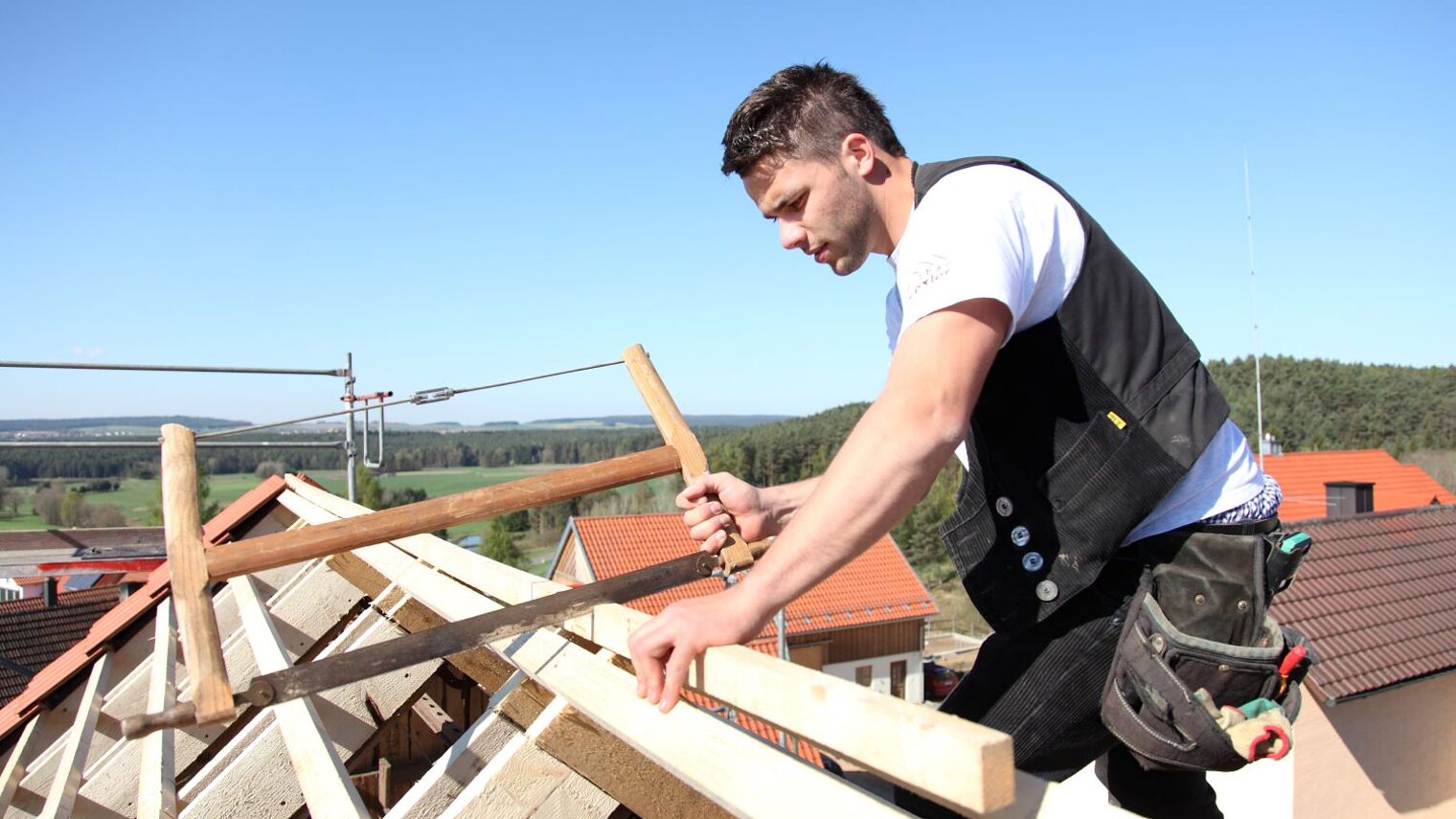 Ein junger Zimmerer mit einer Säge arbeitet auf dem Dach an einem Dachstuhl.