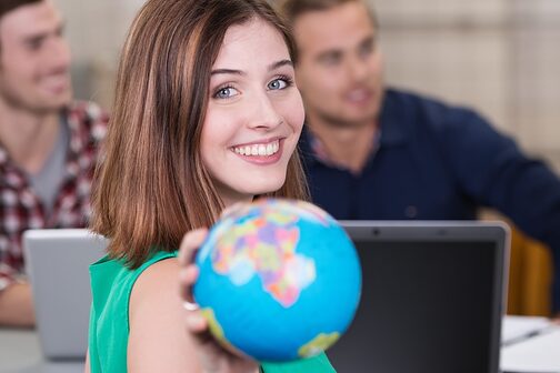 Eine junge Frau sitzt mit weiteren Mitschülern in einem Klassenraum oder Seminarraum und dreht sich zur lächelnd zur Kamera, in der Hand einen kleinen Globus.
