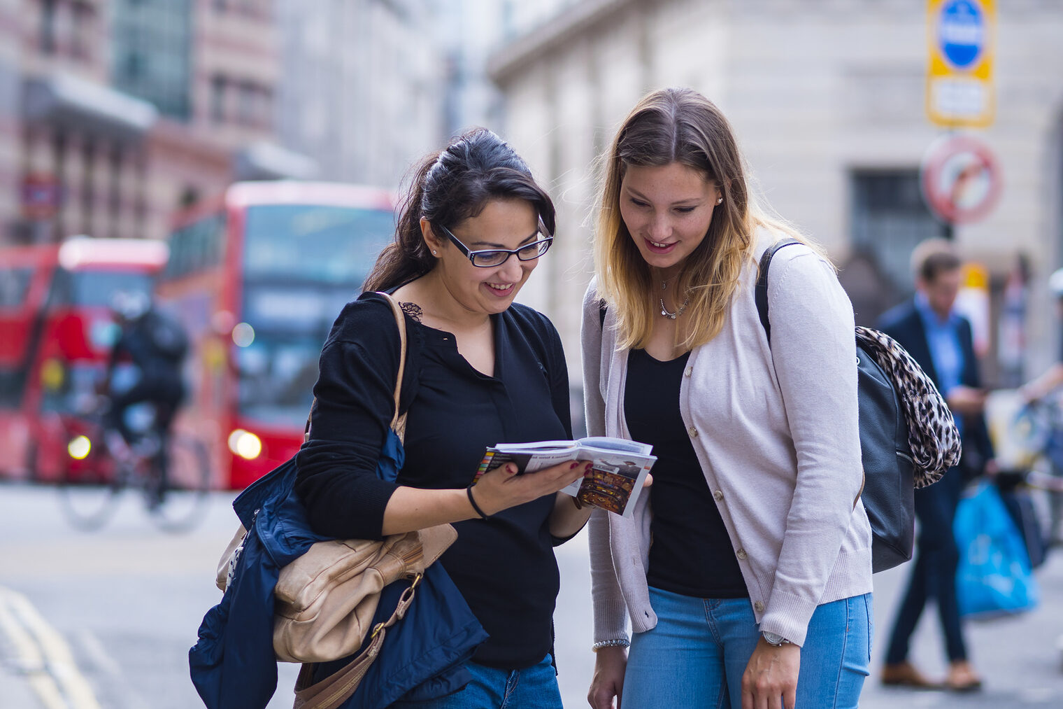 Zwei junge Frauen beugen sich auf einer Straße über einen Stadtplan. Im Hintergrund die für London typischen Busse.