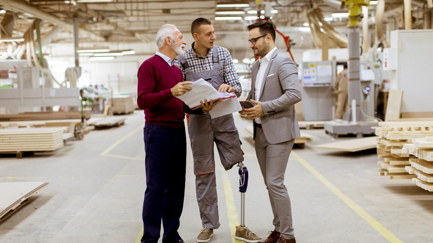 Ein Handwerker mit Beinprothese unterhält sich in seiner Schreinerei mit 2 weiteren männlichen Personen.