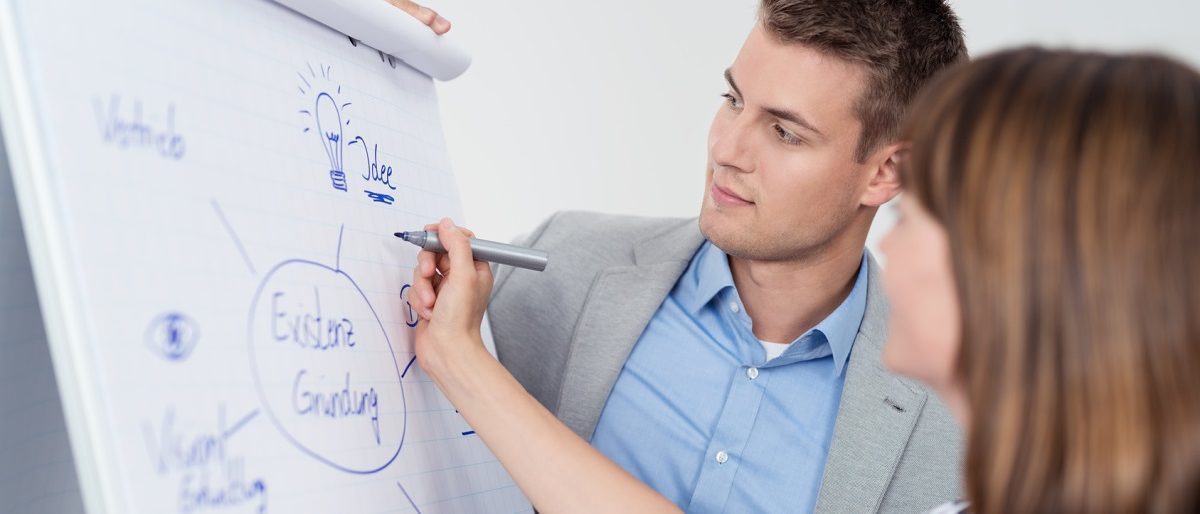 Seminar zur Existenzgründung. Flipchart, auf dem eine Frau etwas zur Existenzgründung schreibt. Seminarleiter schaut zu.