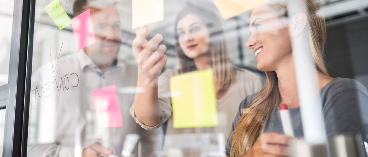 Workshop zum Businessplan. Auf einer Wand kleben Haftzettel mit Ideen. 3 Personen diskutieren.