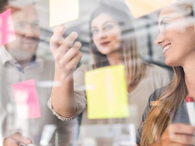 Workshop zum Businessplan. Auf einer Wand kleben Haftzettel mit Ideen. 3 Personen diskutieren.