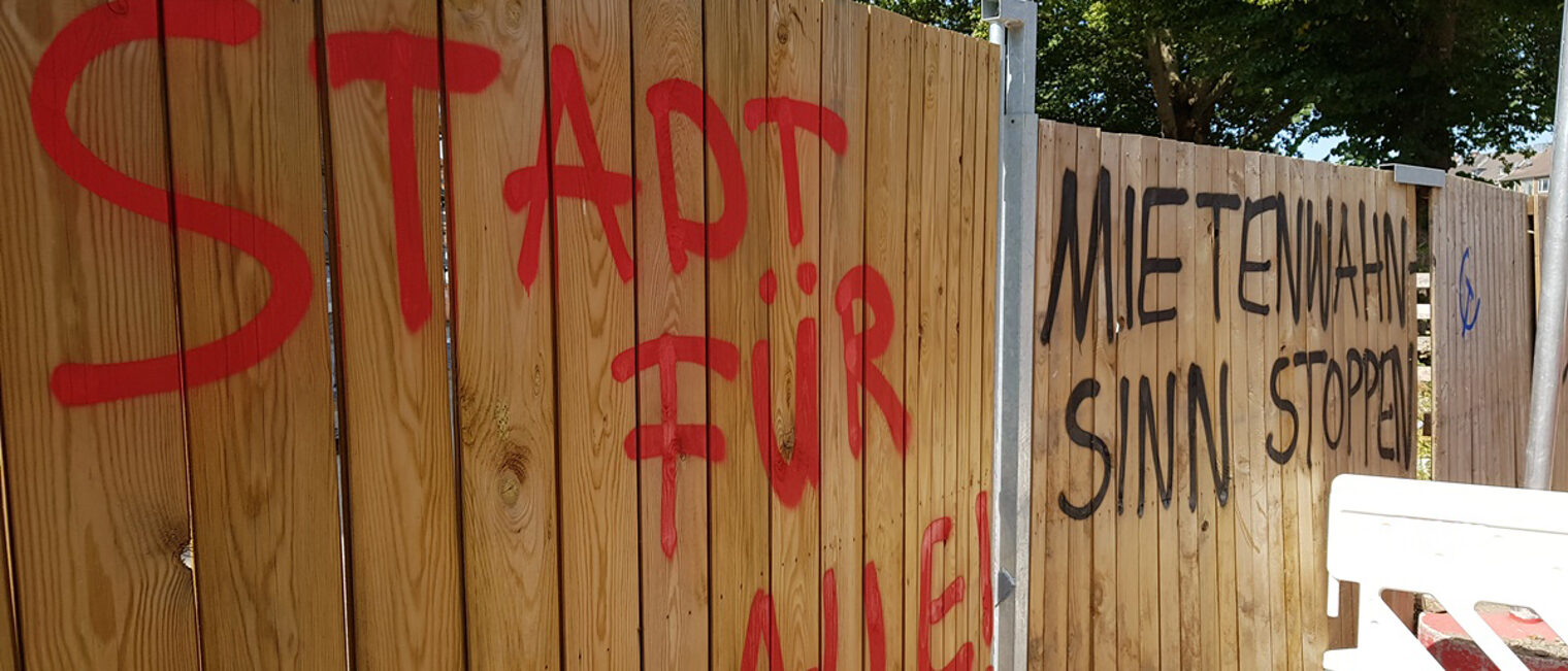 Auf einem Holzbauzaun steht mit gesprühter Farbe "Stadt für Alle" und "Mietwahnsinn stoppen".