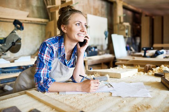 Tischlerin in der Werkstatt telefoniert mit Handy und notiert sich etwas dabei.