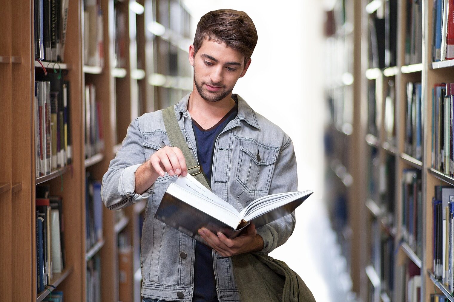 Stundet steht zwischen Bücherregalen in Bibliothek und liest in Buch. 