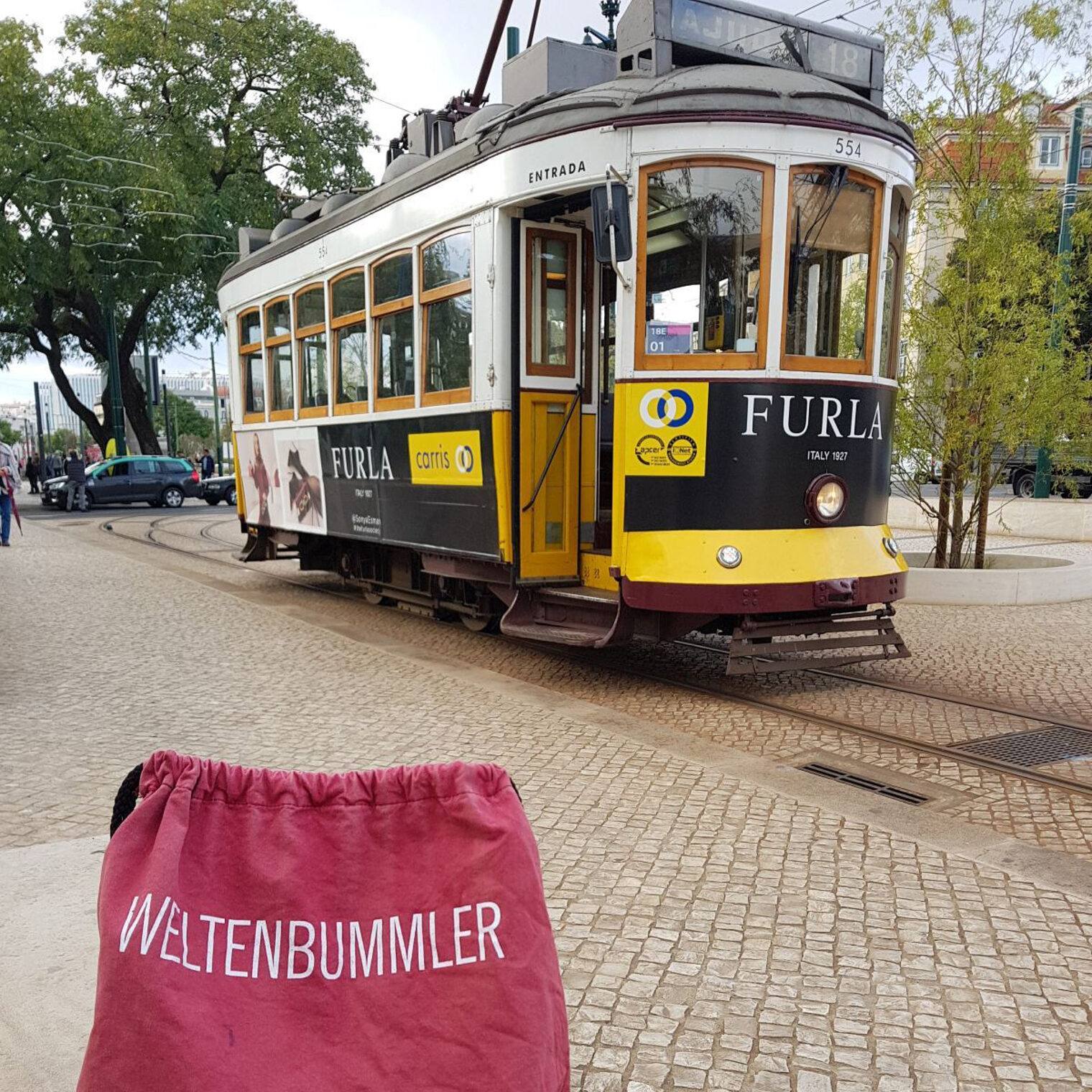 Eine alte Straßenbahn auf einem Platz in Lissabon, davor ein kleiner Rucksack mit der Aufschrift Weltenbummler.
