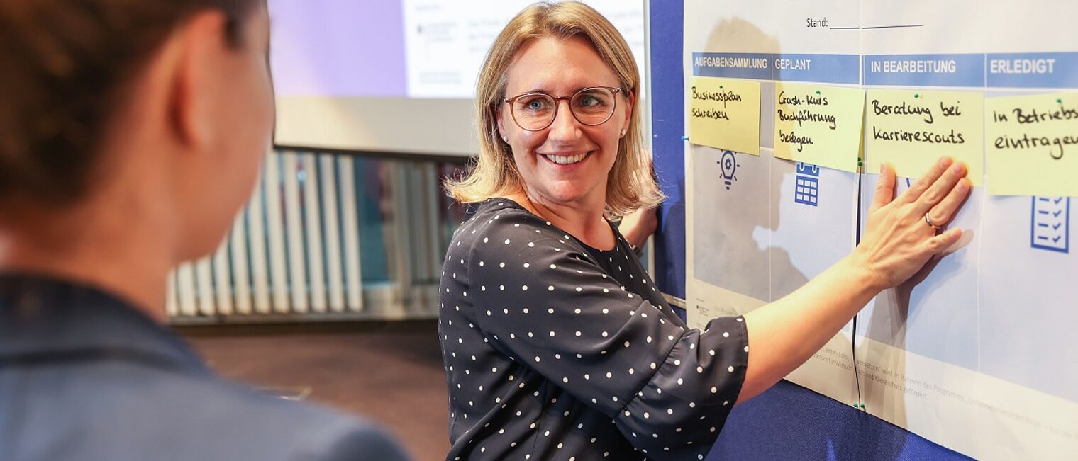 Rebecca Haag Karrierescout Rebecca Haag steht an einer Pinn-Board mit Post-its und erklärt einer Kursteilnehmerin die Schritte zur Selbständigkeit.