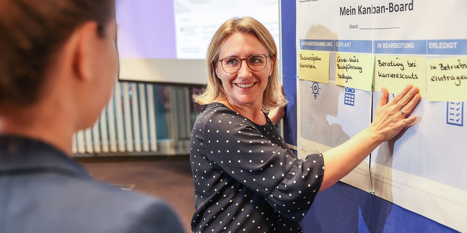 Karrierescout Rebecca Haag steht an einer Pinn-Board mit Post-its und erklärt einer Kursteilnehmerin die Schritte zur Selbständigkeit.