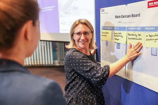 Karrierescout Rebecca Haag steht an einer Pinn-Board mit Post-its und erklärt einer Kursteilnehmerin die Schritte zur Selbständigkeit.