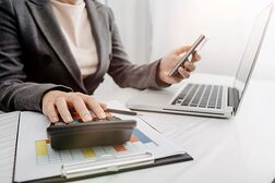 Eine weibliche Person am Schreibtisch sitzend arbeitet mit Laptop und Taschenrechner.
