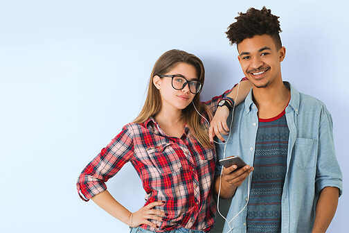 Jugendliches Mädchen und Junge hören über Kopfhörer Musik und schauen freundlich in die Kamera.