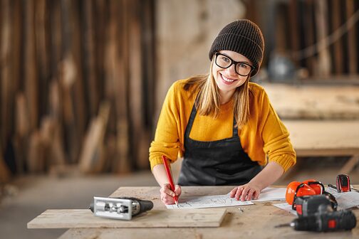 Fröhliche junge Tischlerin, die in die Kamera schaut, während sie Dokumente prüft und in der Handwerkswerkstatt arbeitet