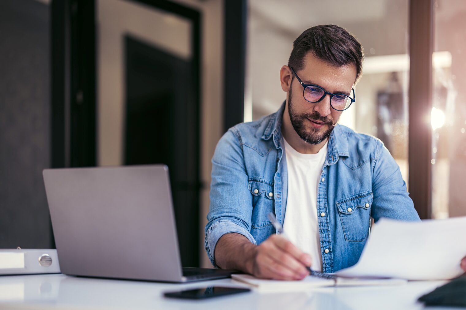 Junger Mann mit Brille, der mit einem Laptop am Schreibtisch sitzt und Papierunterlagen prüft.