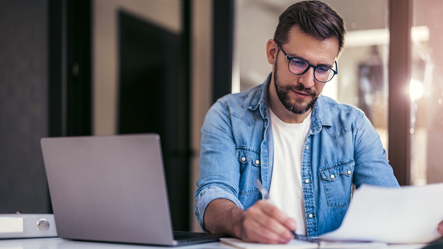 Junger Mann mit Brille, der mit einem Laptop am Schreibtisch sitzt und Papierunterlagen prüft.