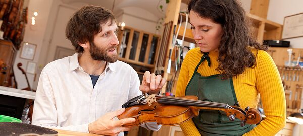 Geigenbauer erklärt einer jungen Frau in einer Werkstatt die Bauteile einer Geige