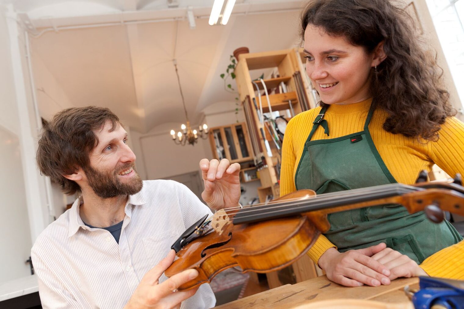 Geigenbauer erklärt einer jungen Frau in einer Werkstatt die Bauteile einer Geige