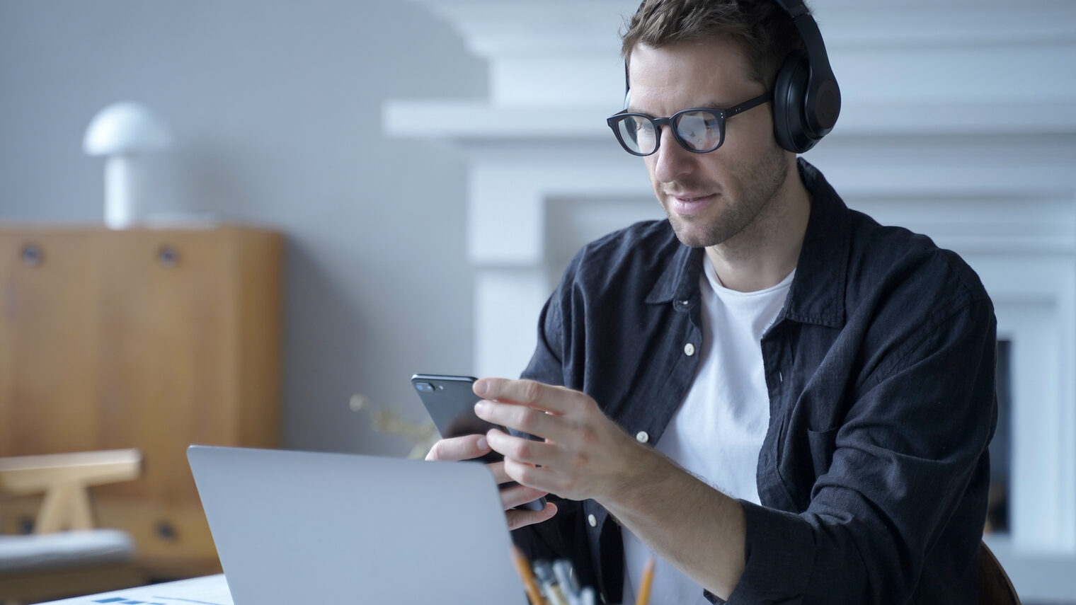 Männlicher Unternehmer mit Kopfhörern, der am Schreibtisch im Home Office sitzt und das Mobiltelefon benutzt