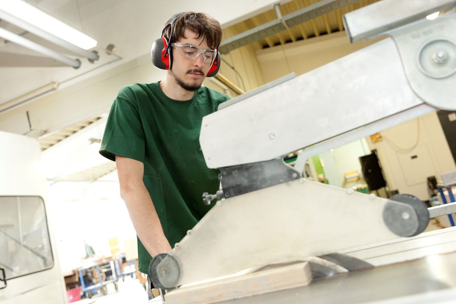 Junger Mann mit Kopfschutzhörern arbeitet in einer Tischlerei an der Säge.