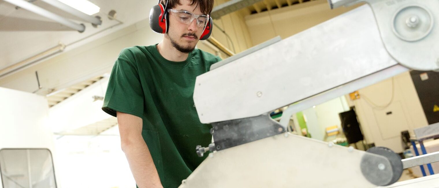 Junger Mann mit Kopfschutzhörern arbeitet in einer Tischlerei an der Säge.