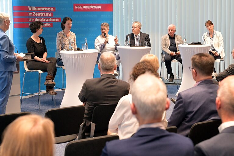 Podiumsdiskussion mit Moderatorin und 6 Teilnehmenden an Stehtischen vor Saalpublikum