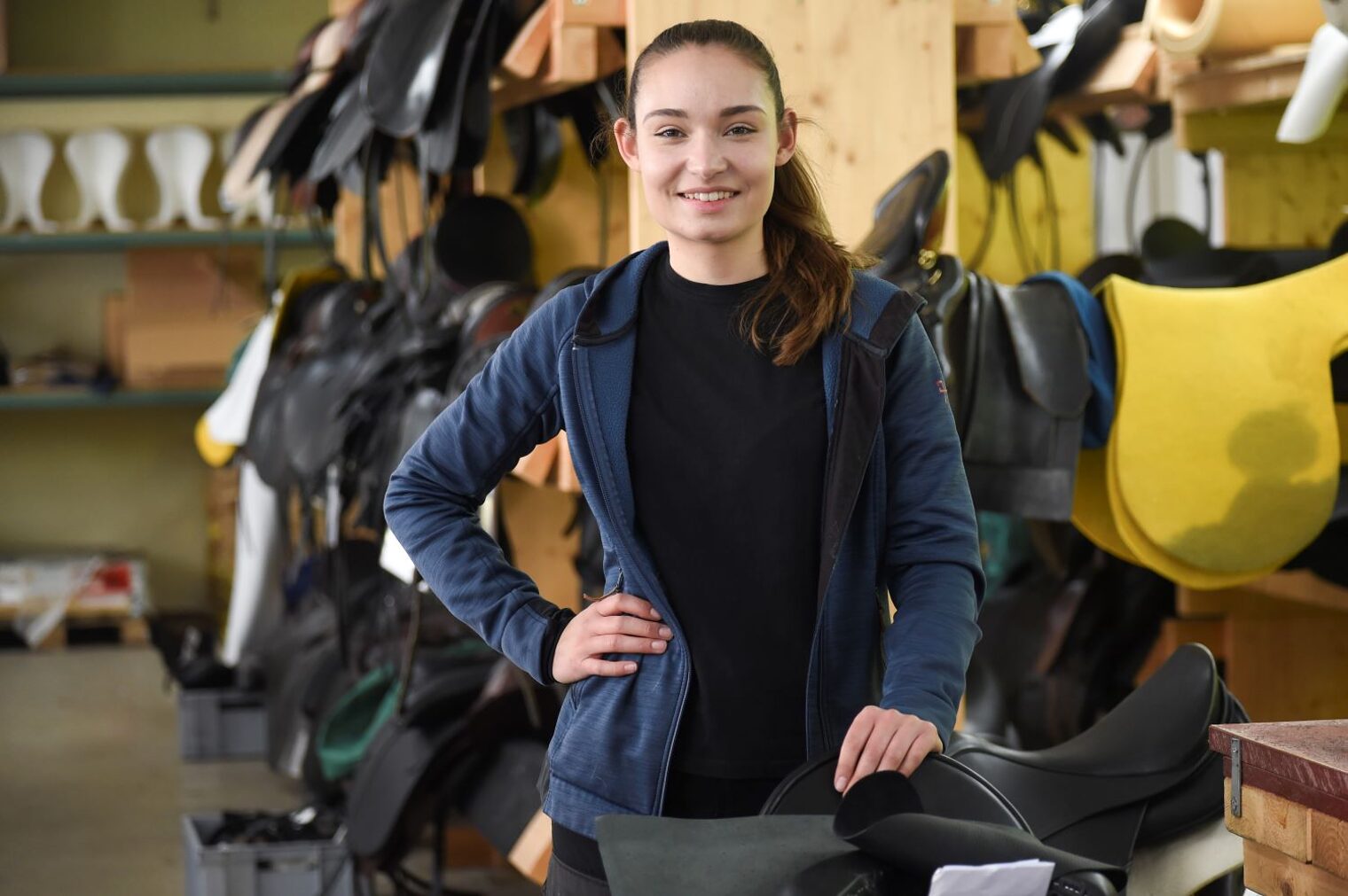 Alexandra Funk steht in einer Sattler-Werkstatt. Im Hintergrund verschiedene Reitsättel.