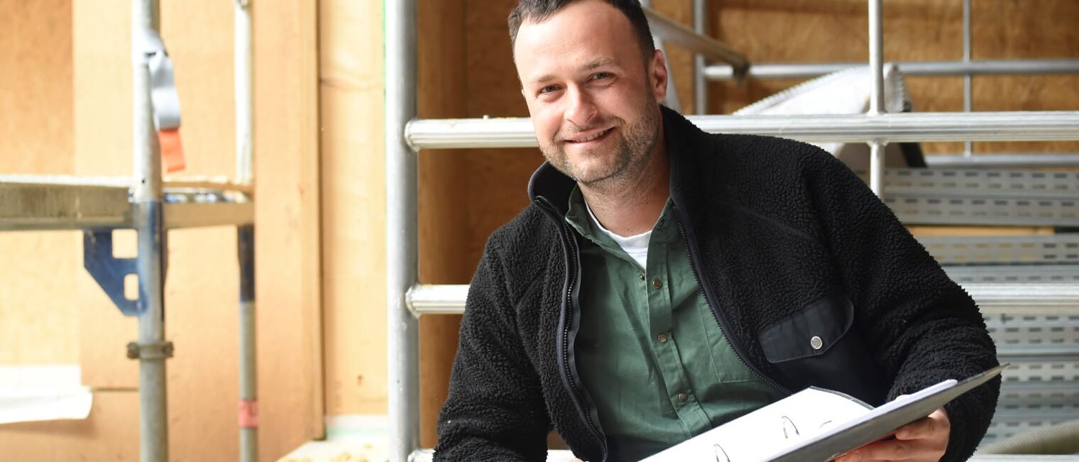 Anton Rößger sitzt auf einer Baustelle in der Hocke und hält einen Aktenordner in der Hand.