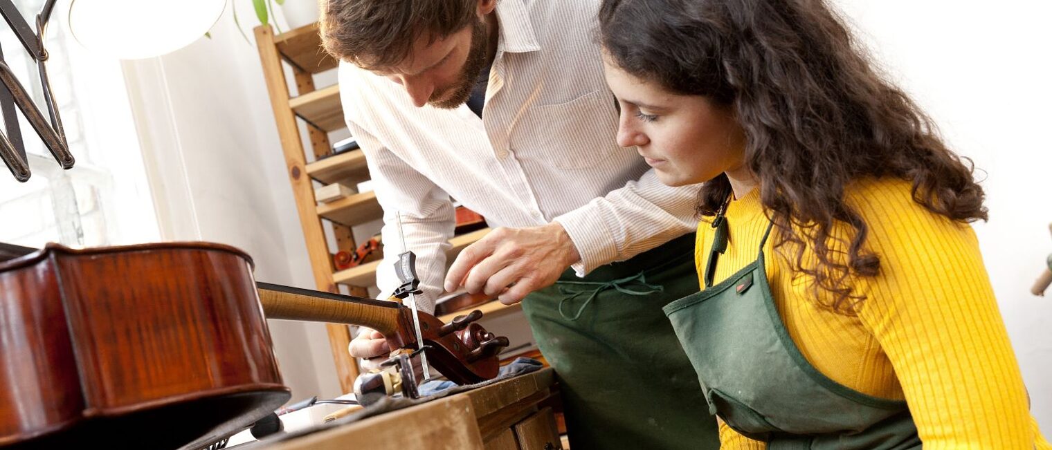 Ein Geigenbauer in seiner Werkstatt zeigt einer jungen Frau, wie man an einer Geige arbeitet.