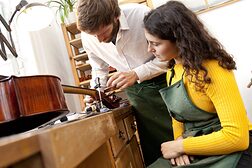 Ein Geigenbauer in seiner Werkstatt zeigt einer jungen Frau, wie man an einer Geige arbeitet.