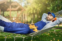 Männlicher Handwerker in Overall, der an einem sonnigen Tag mit geschlossenen Augen in einer Hängematte im Freien liegt.
