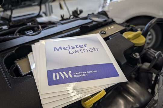 Mehrere Aufkleber mit Text "Meisterbetrieb" und Logo Handwerkskammer Düsseldorf liegen auf einem PKW-Motorblock.