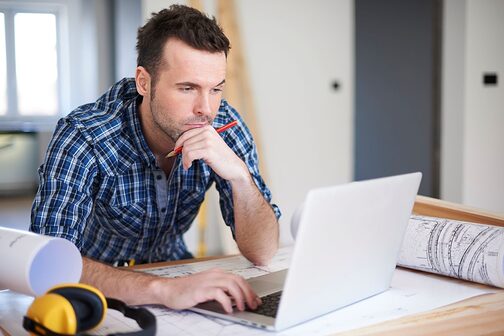 Mann am Schreibtisch sitzt nachdenklich vor seinem Laptop. Rechts und links von ihm legen Bauzeichnungen auf dem Schreibtisch.