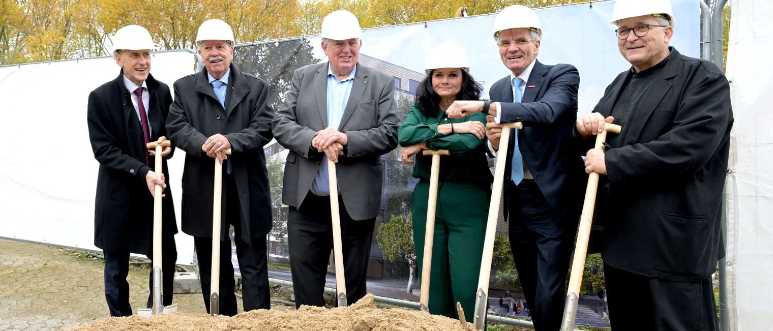 6 Personen aus HWK-Führungspitze und NRW-Ministerium posieren mit ihren Spaten an der Baustelle.