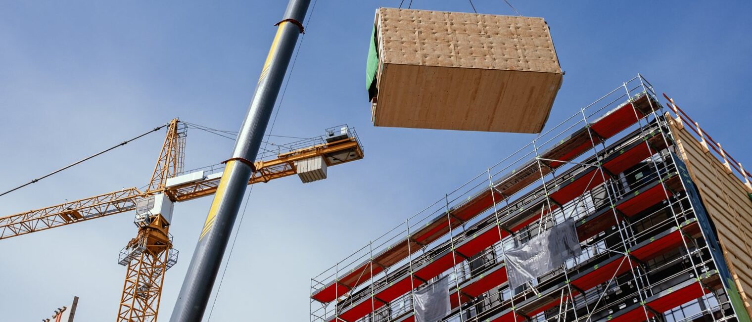 Ein Baustellenkran hebt ein vorgefertigtes Holzbaumodul an seine Position.
