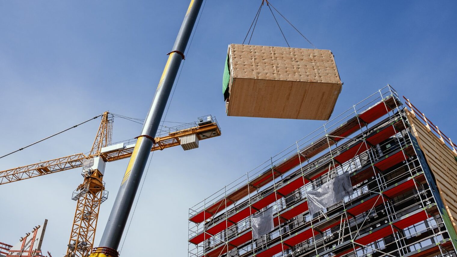 Ein Baustellenkran hebt ein vorgefertigtes Holzbaumodul an seine Position.