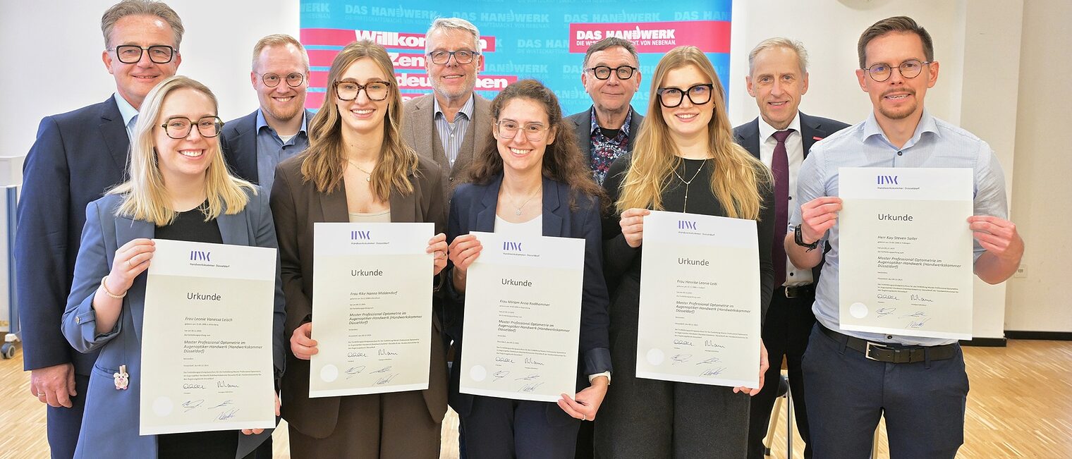 Die Absolvierenden posieren für ein Foto. Sie zeigen ihre Urkunden. Im Bildhintergrund sind Handwerksfunktionäre zu sehen.