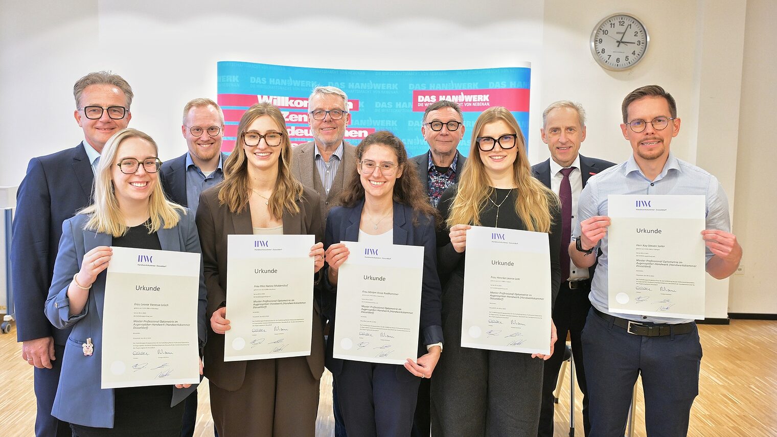 Die Absolvierenden posieren für ein Foto. Sie zeigen ihre Urkunden. Im Bildhintergrund sind Handwerksfunktionäre zu sehen.
