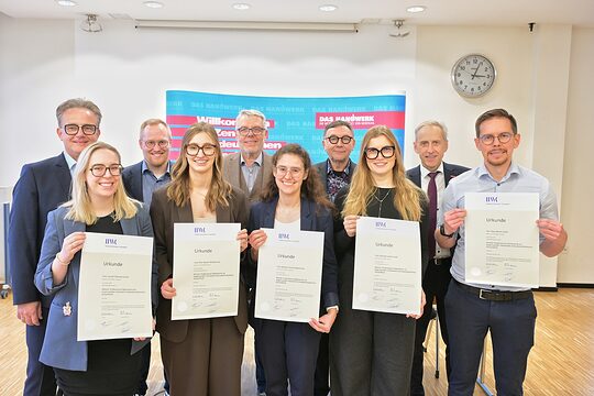 Die Absolvierenden posieren für ein Foto. Sie zeigen ihre Urkunden. Im Bildhintergrund sind Handwerksfunktionäre zu sehen.
