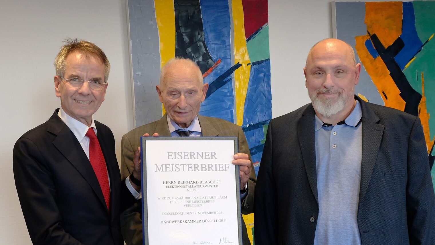 Reinhard Blaschke zeigt seinen Eisernen Meisterbrief. Er posiert mit zwei Gratulanten vor zwei bunten Bildern.