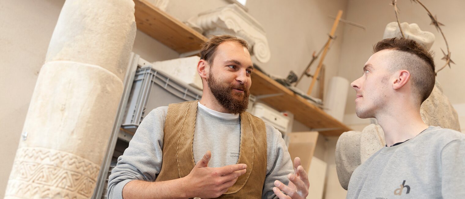 Steinmetz-Ausbilder spricht mit Azubi in Werkstatt.