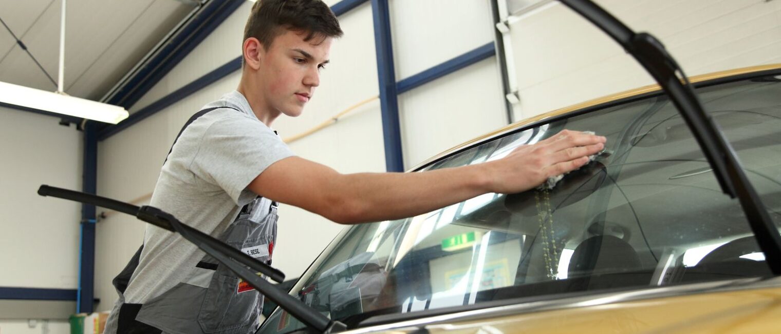 Ein junger Mann wischt in einer Autowerkstatt über die Frontscheibe eines Fahrzeugs