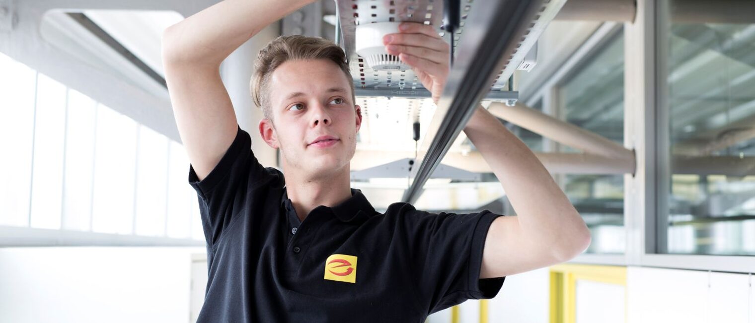 Ein junger Informationselektroniker arbeitet kopfüber an einer Deckeninstallation.