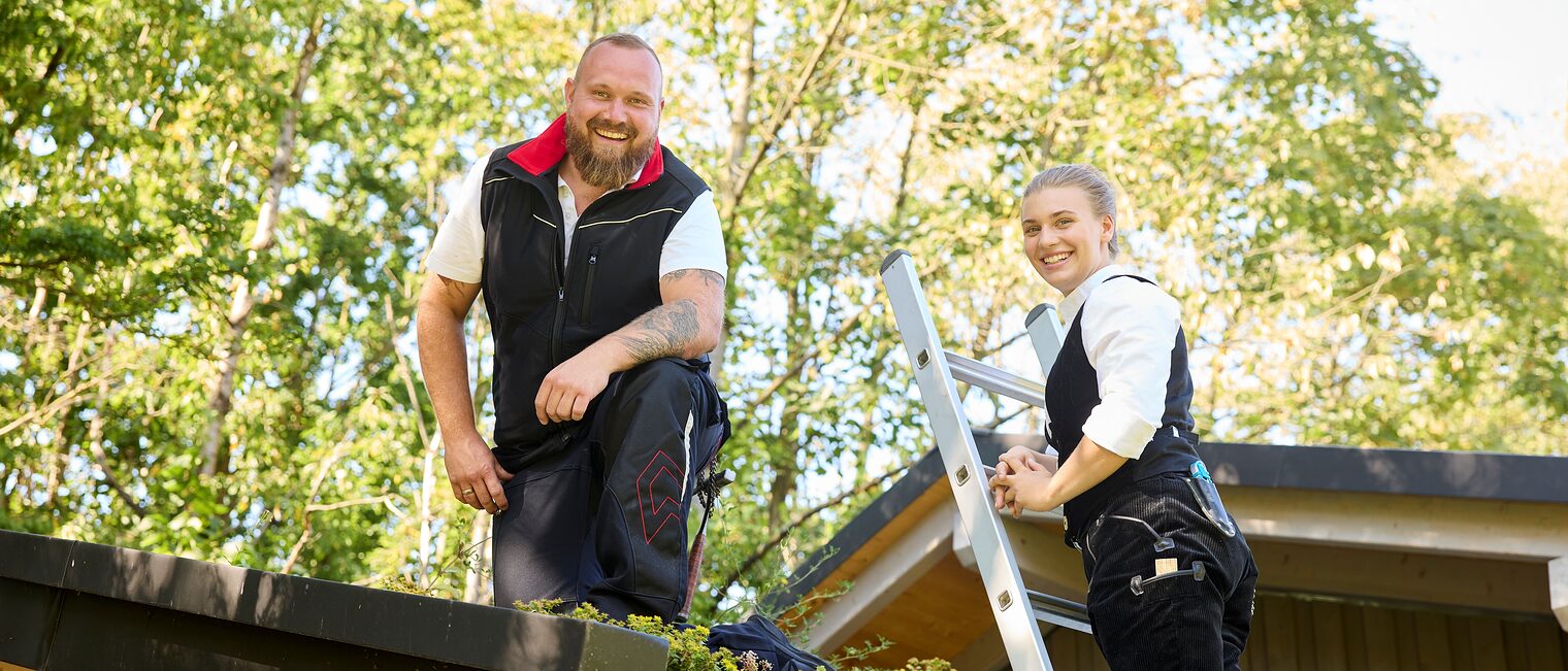 Ein Mann in Arbeitskleidung kniet auf einem Dach, daneben steht eine Frau auf einer Leiter