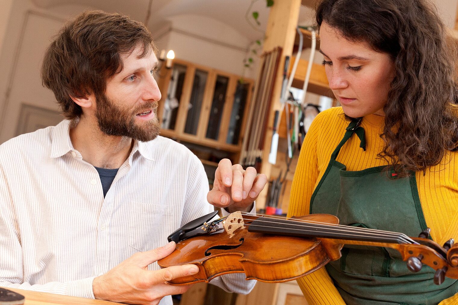 Geigenbauer erklärt Praktikantin in der Werkstatt die Bauteile einer Geige.