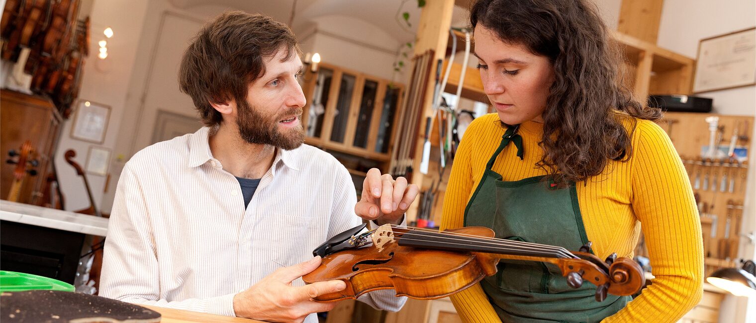 Geigenbauer erklärt Praktikantin in der Werkstatt die Bauteile einer Geige.