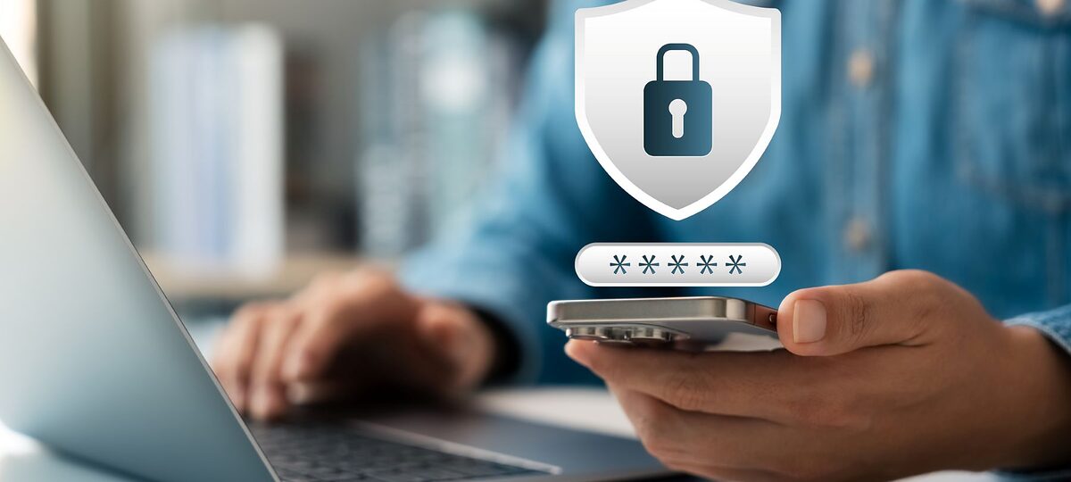 Ein Mann arbeitet am Laptop mit Smartphone in der einen Hand. Eingebunden ist ein Schloss-Symbol für Cybersicherheit.