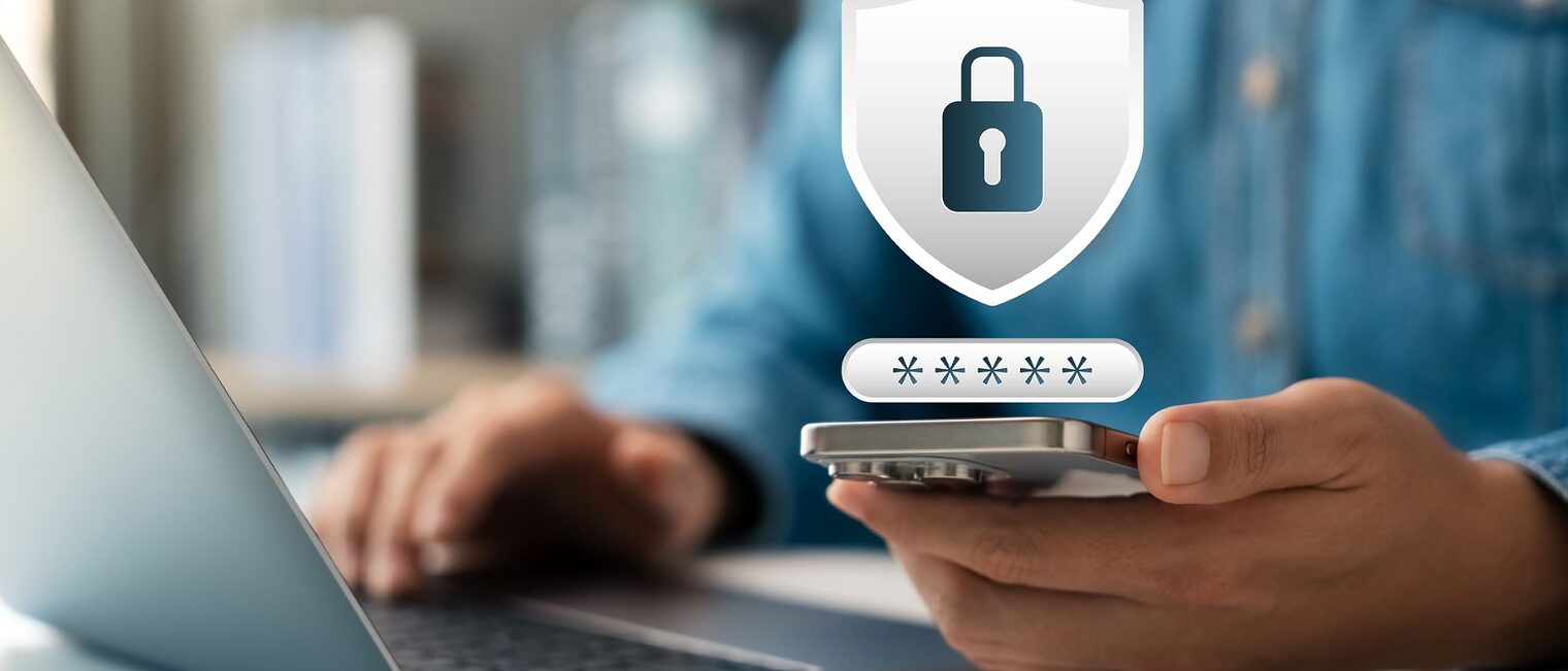 Ein Mann arbeitet am Laptop mit Smartphone in der einen Hand. Eingebunden ist ein Schloss-Symbol für Cybersicherheit.