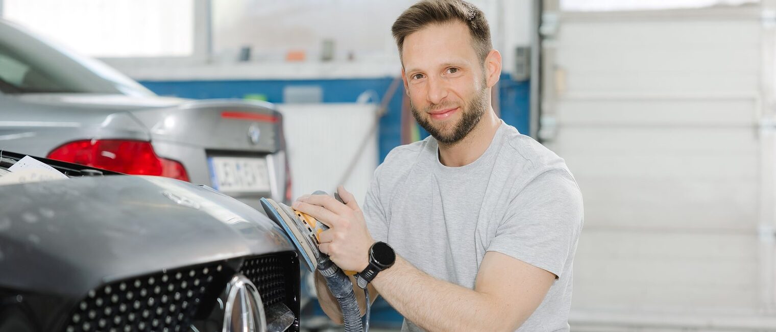 Selim Cakmak hockt vor einem Auto und schleift an der Kühlerhaube. Er blickt in die Kamera.