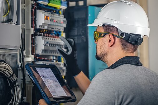 Ein Elektrotechniker oder Installateur arbeitet mit einem Tablet an einer Schalteinheit mit Sicherungen.