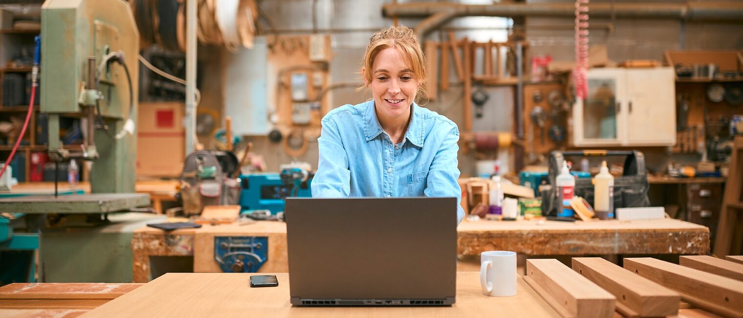 Eine Tischlerin arbeitet an einem Laptop in einer Holzwerkstatt. 